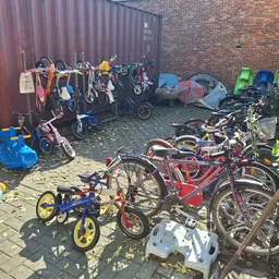 Verzameling tweedehands fietsen, kinderfietsjes en speelgoed in een opslagruimte tegen een bakstenen muur