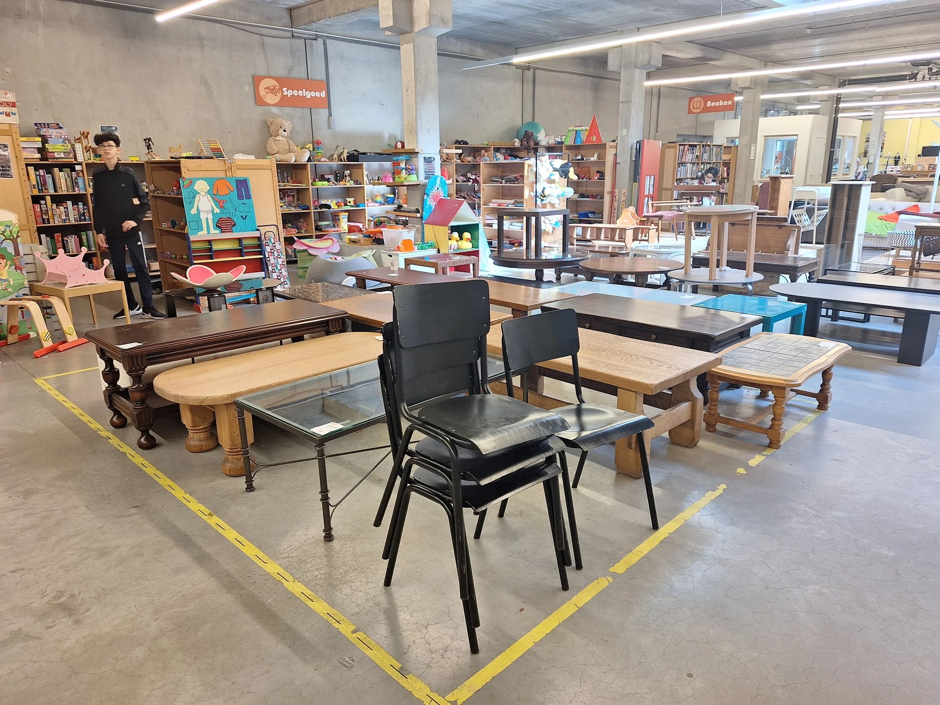Werkruimte met tafels en stoelen in kringloopwinkel, omgeven door boeken en spullen
