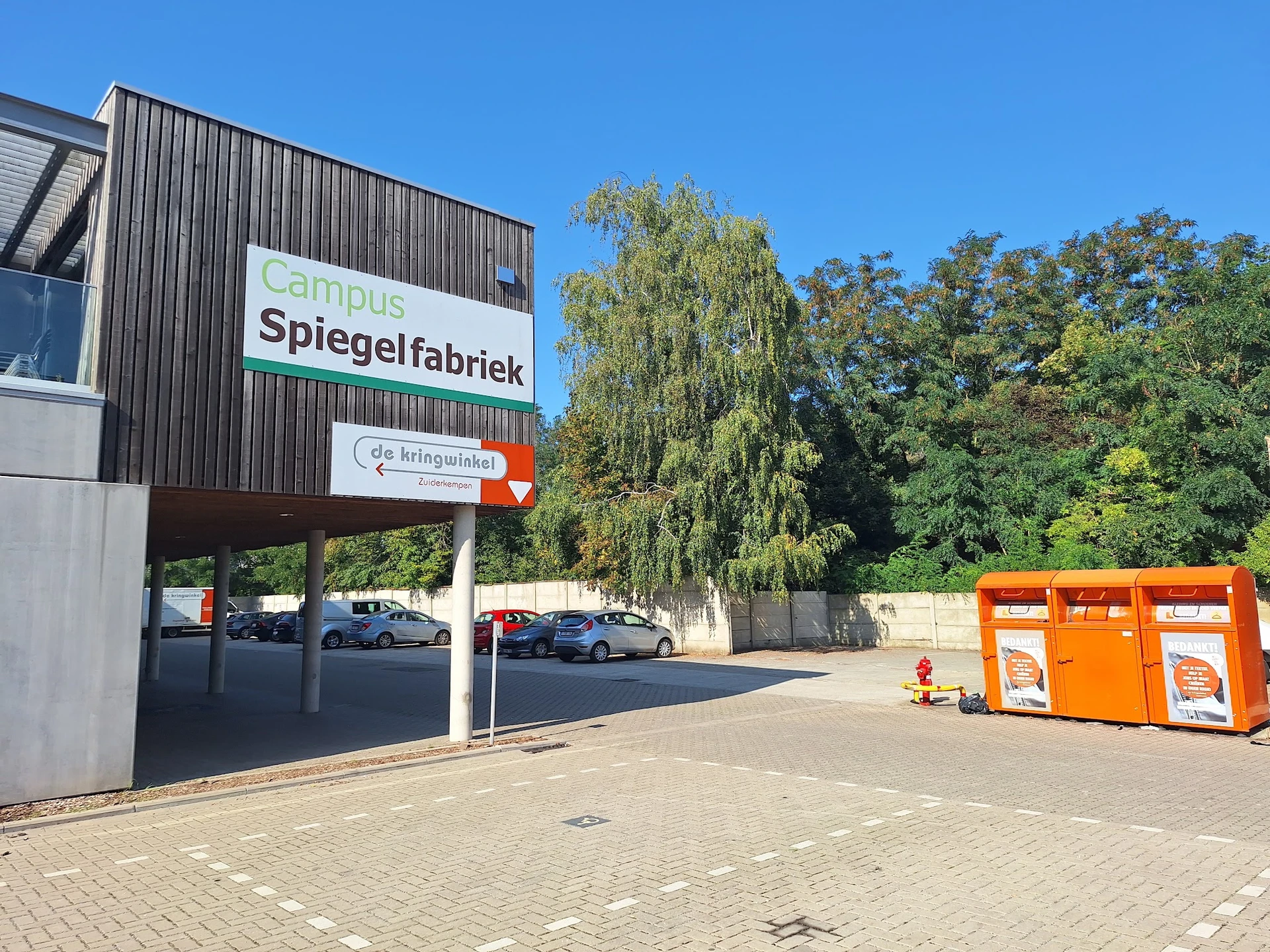 Voorgevel van kringloopwinkel met bord 'Spiegelfabriek', parkeerplaats en oranje containers