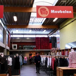 Overzicht van de winkelruimte met kledingrekken, rode Meubelen-bord aan het plafond en winkelafdelingen
