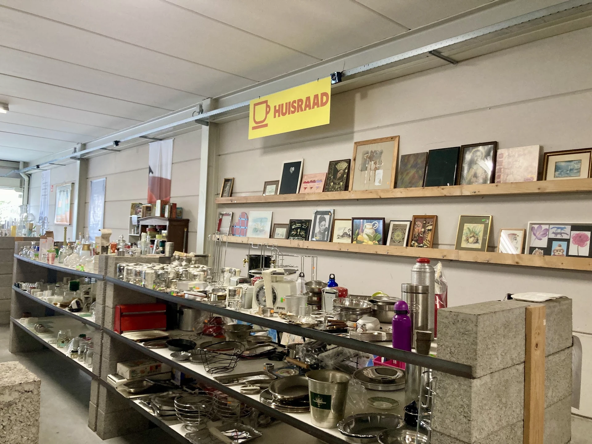 Vitrinekast met huishoudelijke artikelen en serviesgoed in kringloopwinkel, met wandrek met foto's en posters op achtergrond