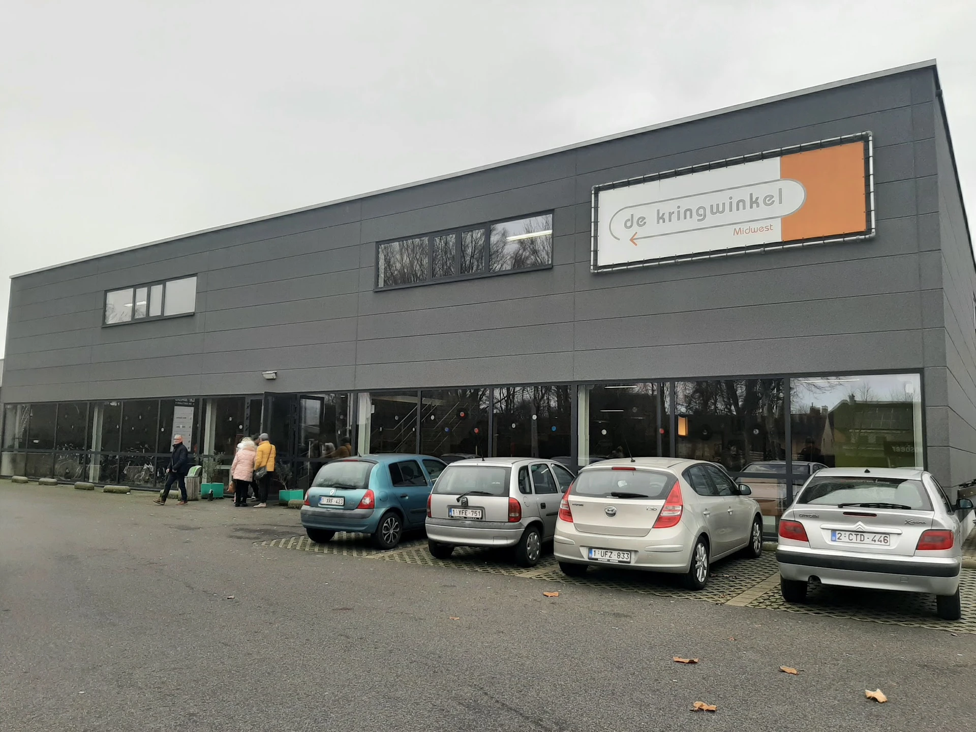 Grijze moderne gevel van kringloopwinkel met grote ramen en oranje logo, parkeerplaats met enkele auto's ervoor