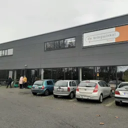 Grijze moderne gevel van kringloopwinkel met grote ramen en oranje logo, parkeerplaats met enkele auto's ervoor