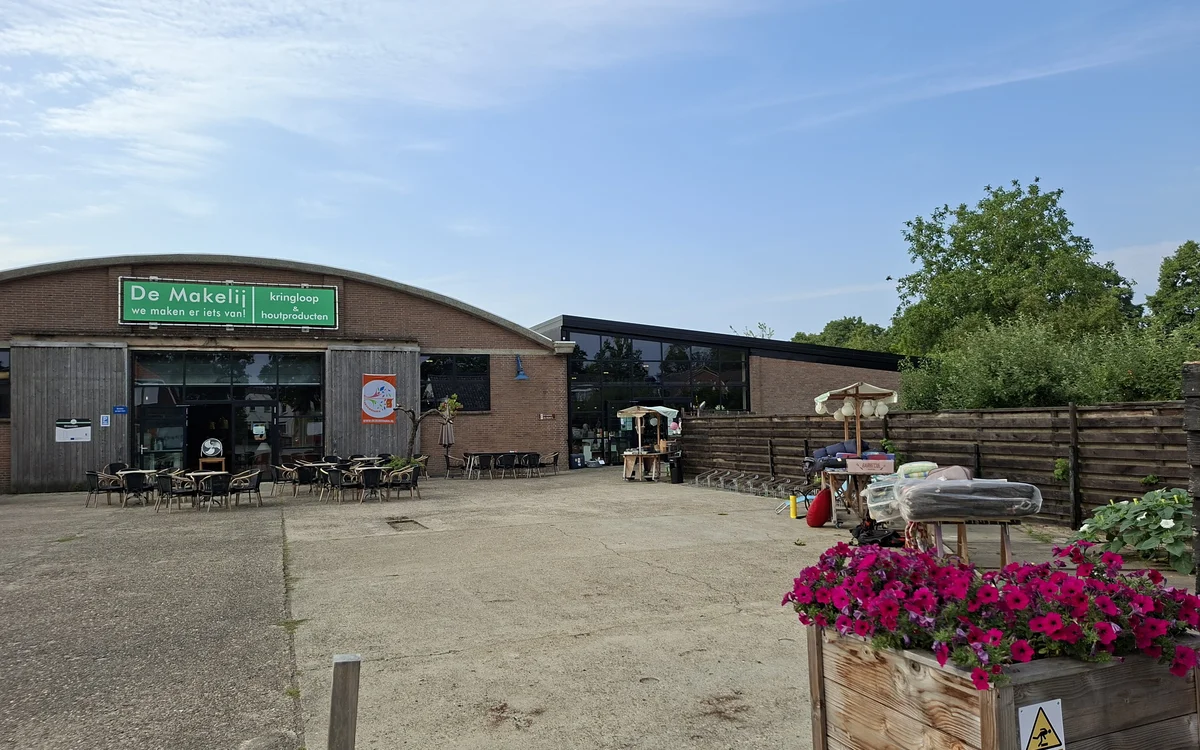 Exterieur van kringloopwinkel De Makelij met groene winkelname op dak, terras met stoelen en bloembakken.