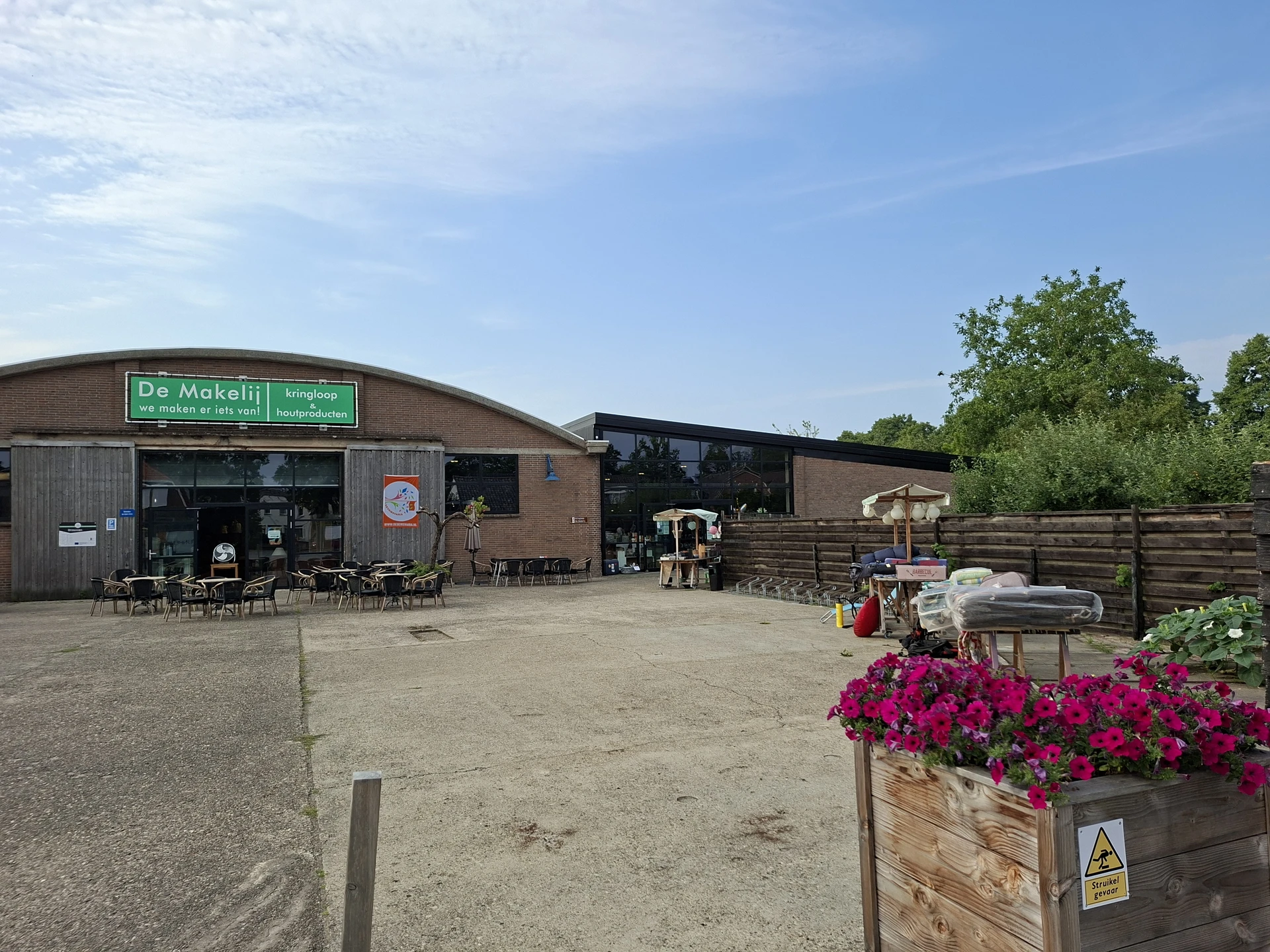 Exterieur van kringloopwinkel De Makelij met groene winkelname op dak, terras met stoelen en bloembakken.