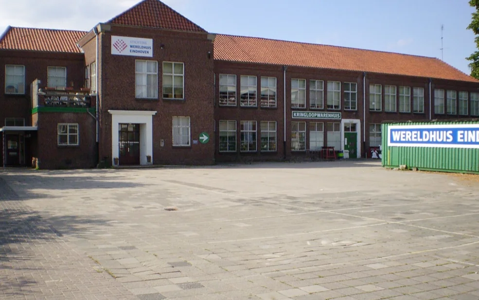 Voorkant van een bakstenen kringloopwinkel met grote ramen en groene accenten, gezien vanaf het plein