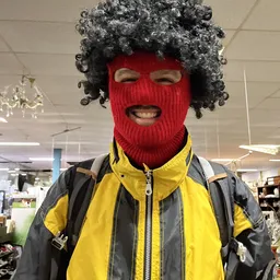 Persoon met grijs krullend haar, rood gebreid masker en geel-zwart jack in een winkelinterieur