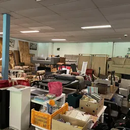 Rommelig winkelinterieur met meubels, dozen, kleding en huishoudelijke artikelen verspreid op de vloer en planken