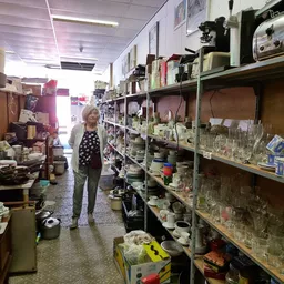 Winkelinterieur met metalen stellingen vol glazen potten, serviesgoed en huishoudspullen; een klant loopt door het middenpad