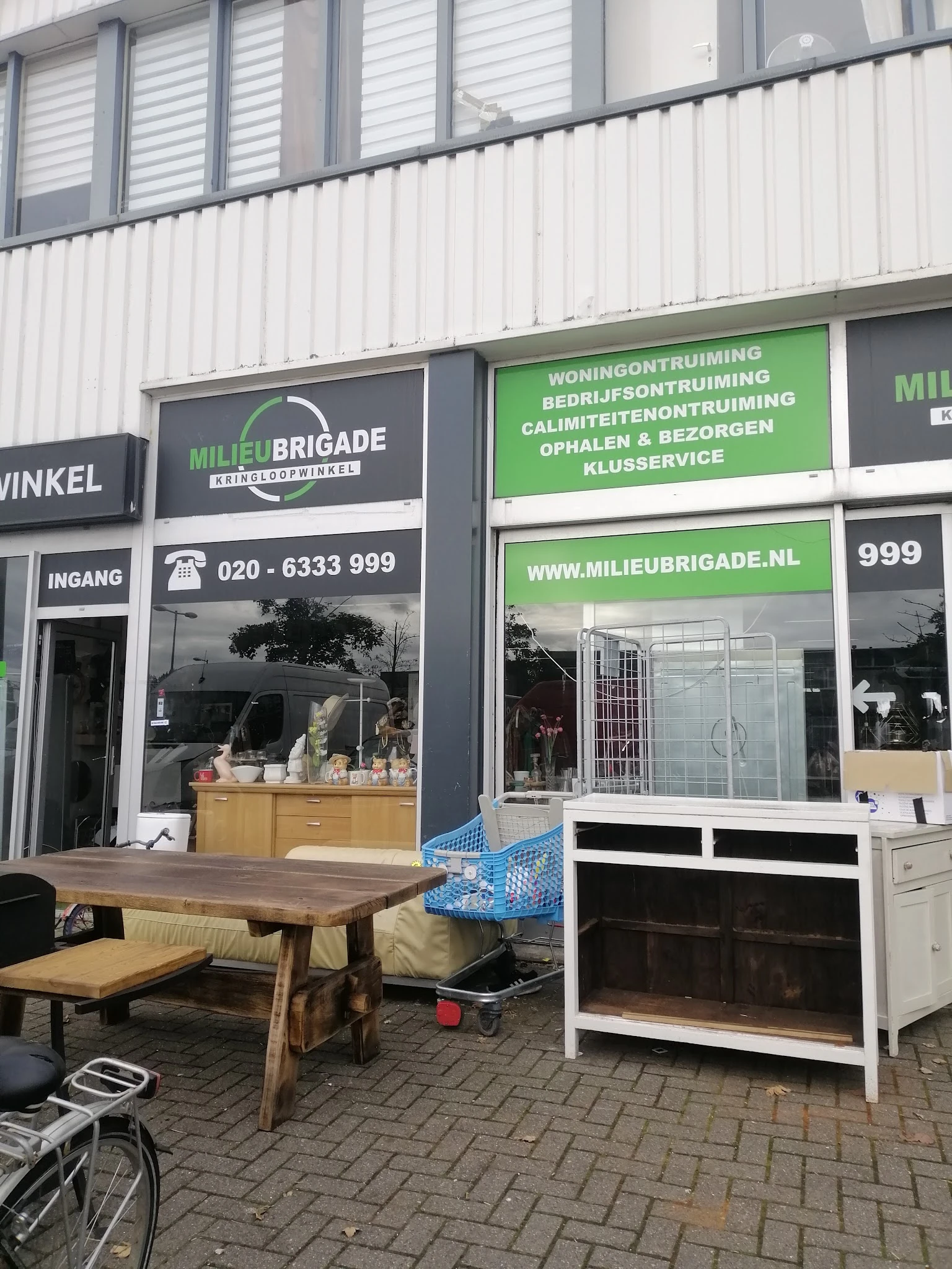 Voorgevel van Milieu Brigade kringloopwinkel met groene en witte belettering, ingang links, ramen met spullen en website-info
