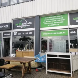 Voorgevel van Milieu Brigade kringloopwinkel met groene en witte belettering, ingang links, ramen met spullen en website-info
