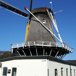 Traditionele Nederlandse windmolen met grijs baksteenwerk en blauwe lucht, tegen een modern wit gebouw.
