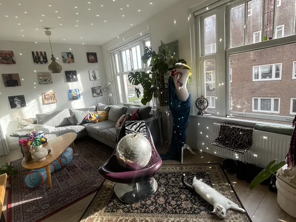 Ruime woonkamer met grijze bank, fauteuil, foto's aan de muur, veel daglicht en planten
