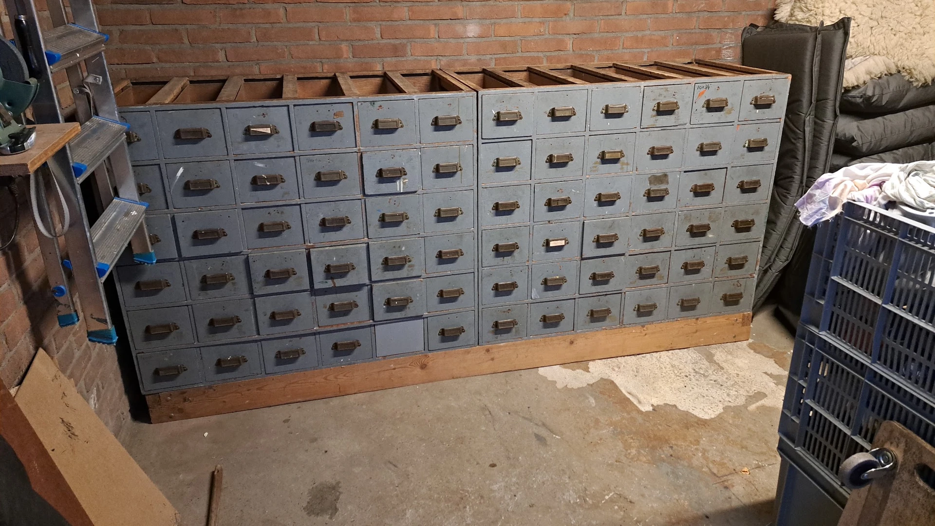Groot kastensysteem met veel grijze laden tegen bakstenen muur, mogelijk archiefkasten of werkbank.