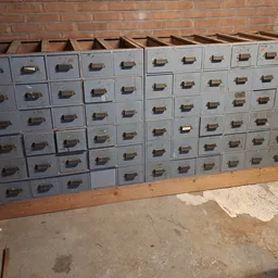 Groot kastensysteem met veel grijze laden tegen bakstenen muur, mogelijk archiefkasten of werkbank.