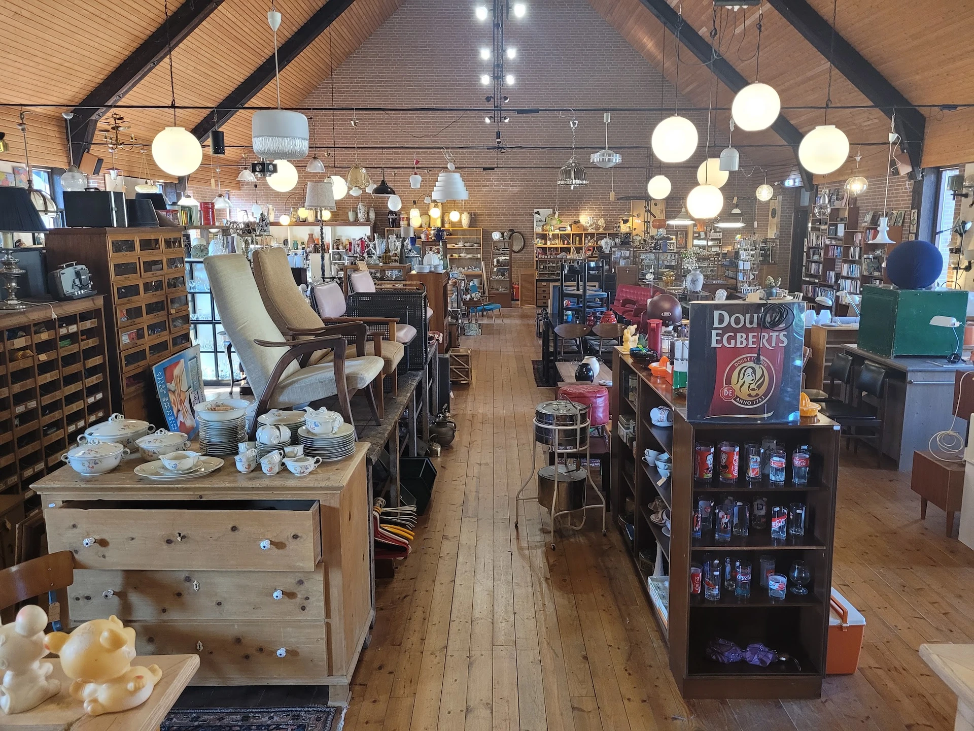 Ruim interieur van kringloopwinkel met houten vloer, stellingen met spullen, ronde hangende lampen en fauteuils op de voorgro
