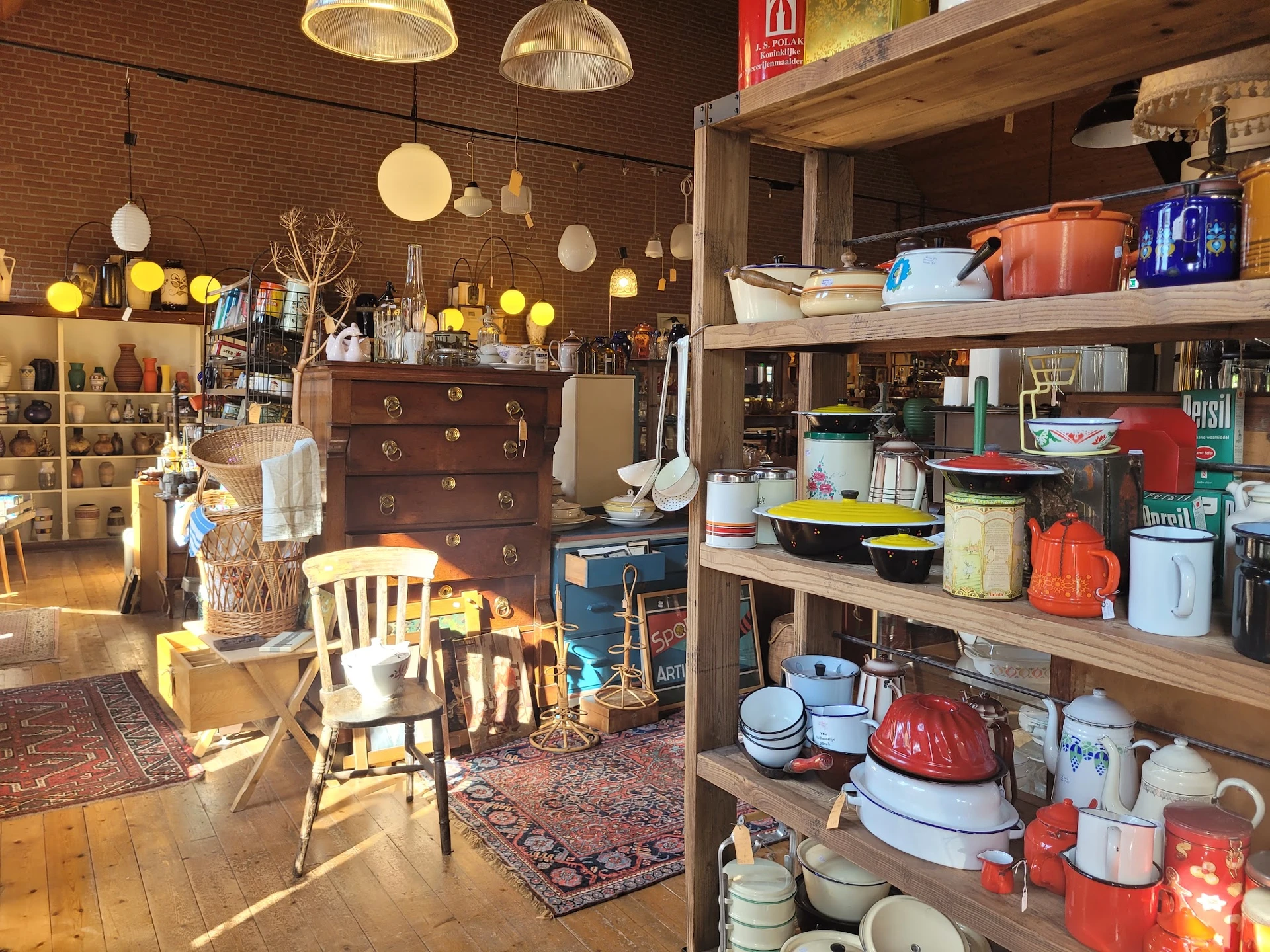 Winkelvloer met houten rekken vol servies, huishoudartikelen, een houten ladenkast, stoelen en diverse spullen onder gloeilam