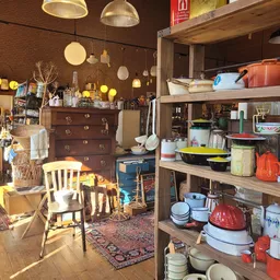 Winkelvloer met houten rekken vol servies, huishoudartikelen, een houten ladenkast, stoelen en diverse spullen onder gloeilam