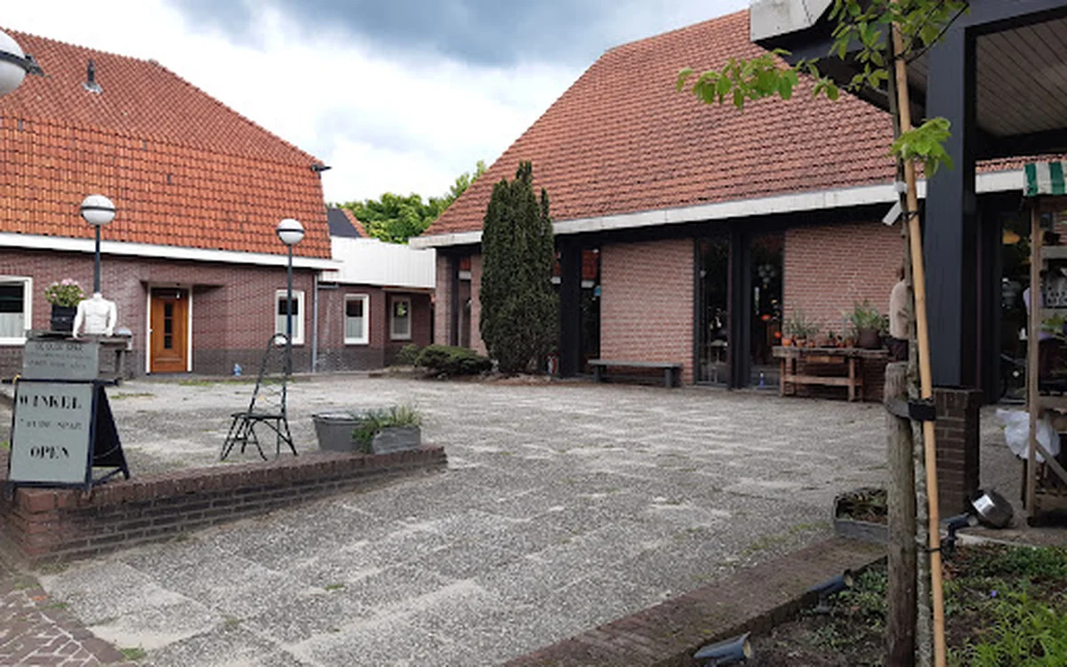 Voorzijde van kringloopwinkel met bakstenen gevels, rode daken en ingang, omgeven door bestrating en groen