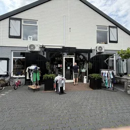 Voorkant van kringloopwinkel met zwarte pui, ramen, uitgestalde kleding op rekken en fietsen voor de ingang