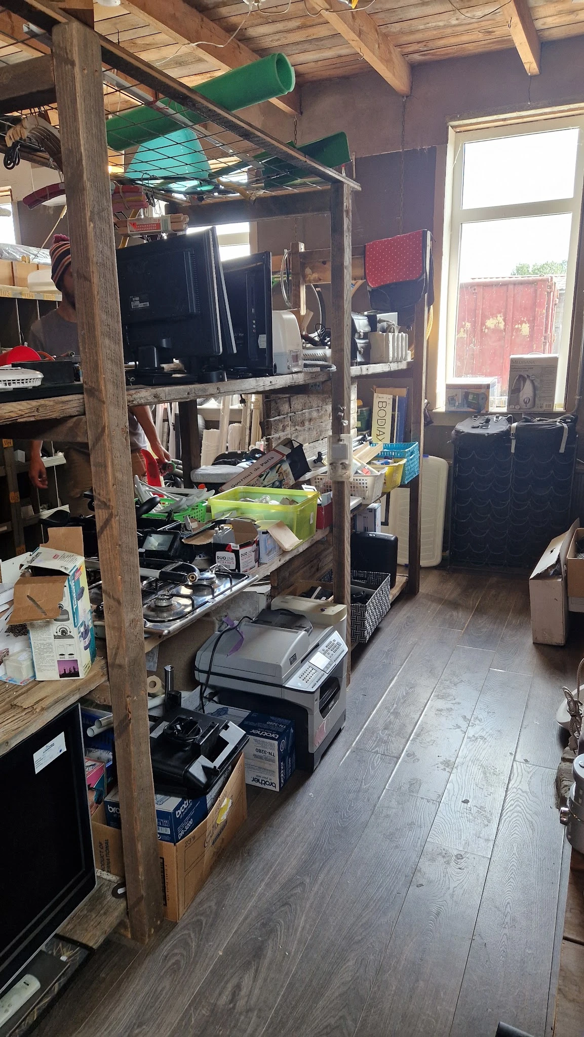 Rommelige winkelvloer met diverse spullen, elektronische apparaten en artikelen verspreid over stellingen en de grond