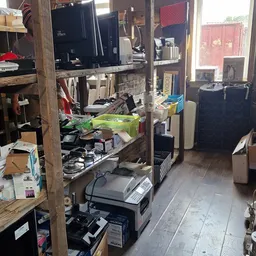 Rommelige winkelvloer met diverse spullen, elektronische apparaten en artikelen verspreid over stellingen en de grond