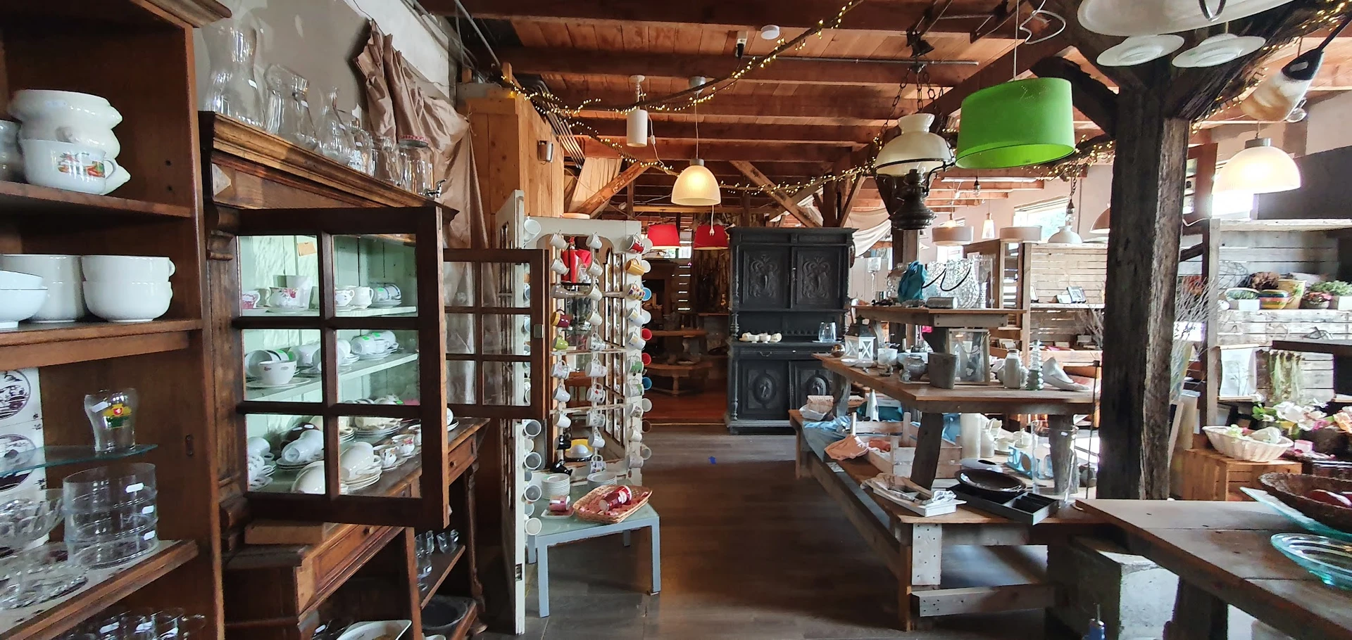 Ruim winkelbinnenland met houten balken, vitrinekastjes met servies, hangende groene lamp en diverse meubelen en spullen