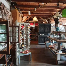 Ruim winkelbinnenland met houten balken, vitrinekastjes met servies, hangende groene lamp en diverse meubelen en spullen