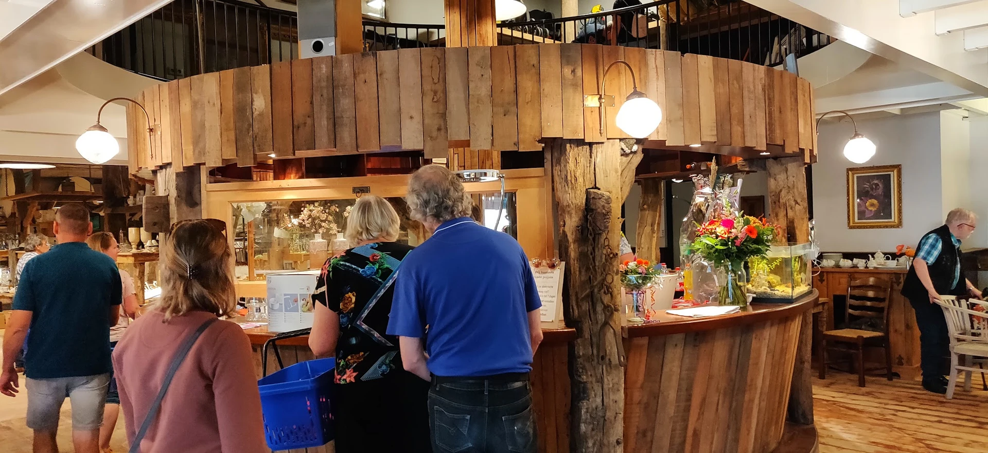 Houten balie van een kringloopwinkel met bloemen en bezoekers eraan, greeploopkasten en houten wanden zichtbaar
