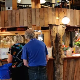 Houten balie van een kringloopwinkel met bloemen en bezoekers eraan, greeploopkasten en houten wanden zichtbaar