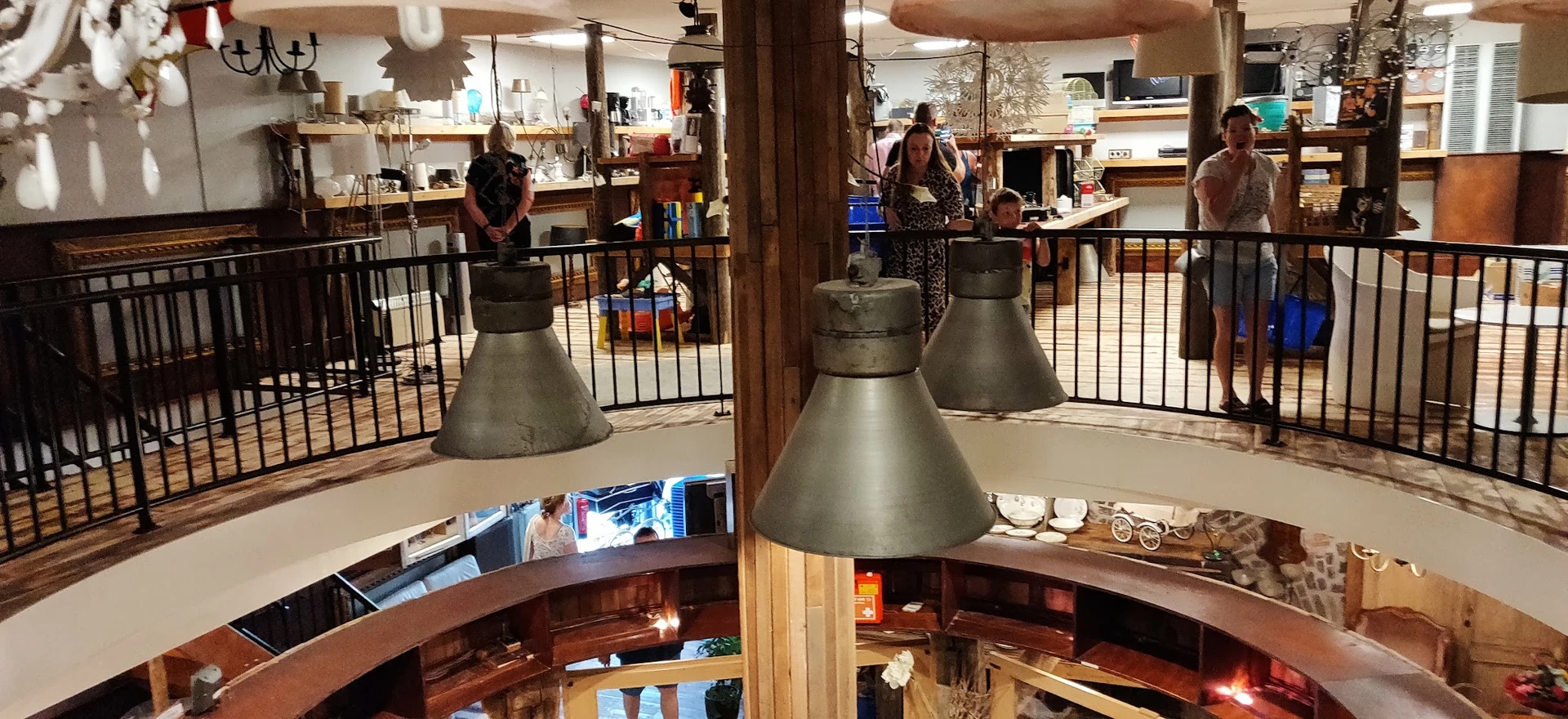 Interieur van kringloopwinkel met hanglampen, zwarte hekwerk, houten balken en bezoekers op verschillende niveaus