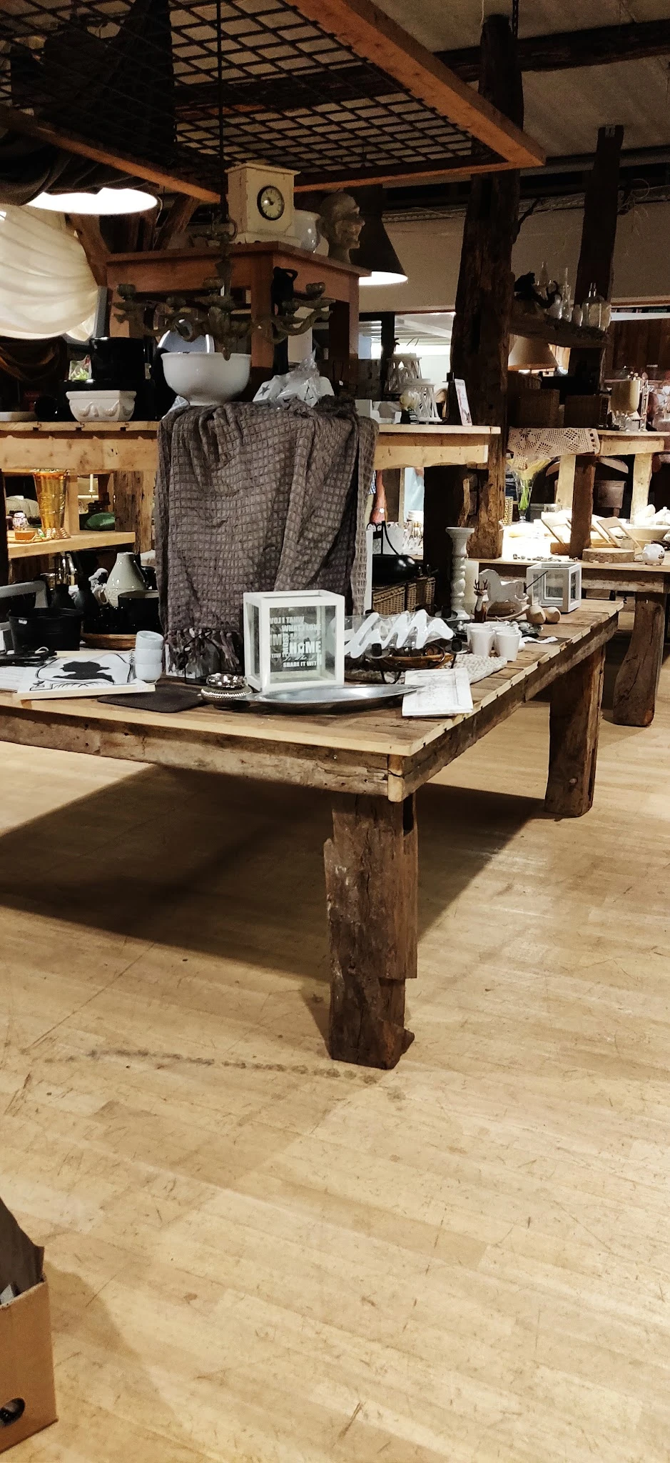 Houten tafel met diverse items zoals witte decoraties, een glazen kast en huishoudelijke spullen in een kringloopwinkel