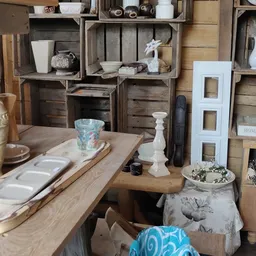 Houten tafel met diverse huishoudartikelen en decoratieve items, tegen achtergrond met houten kisten en stellingen vol spulle