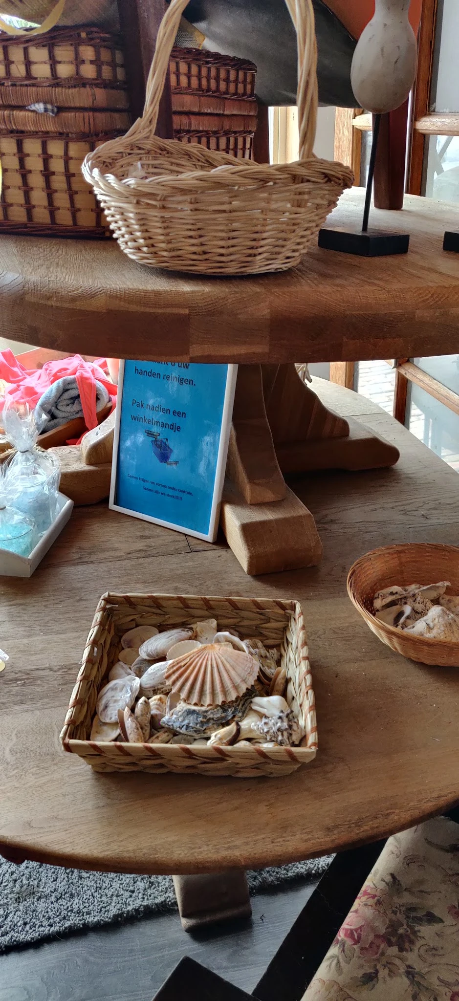 Houten tafeltje met decoratieve items: schelpen in manden, boeken en roze lint in een glas, met zonnlicht van ramen op de ach