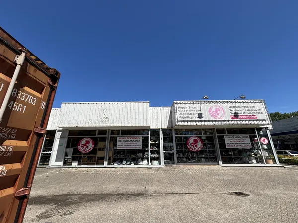 Witte gevel van kringloopwinkel met grote ramen en roze logo's, onder blauwe hemel