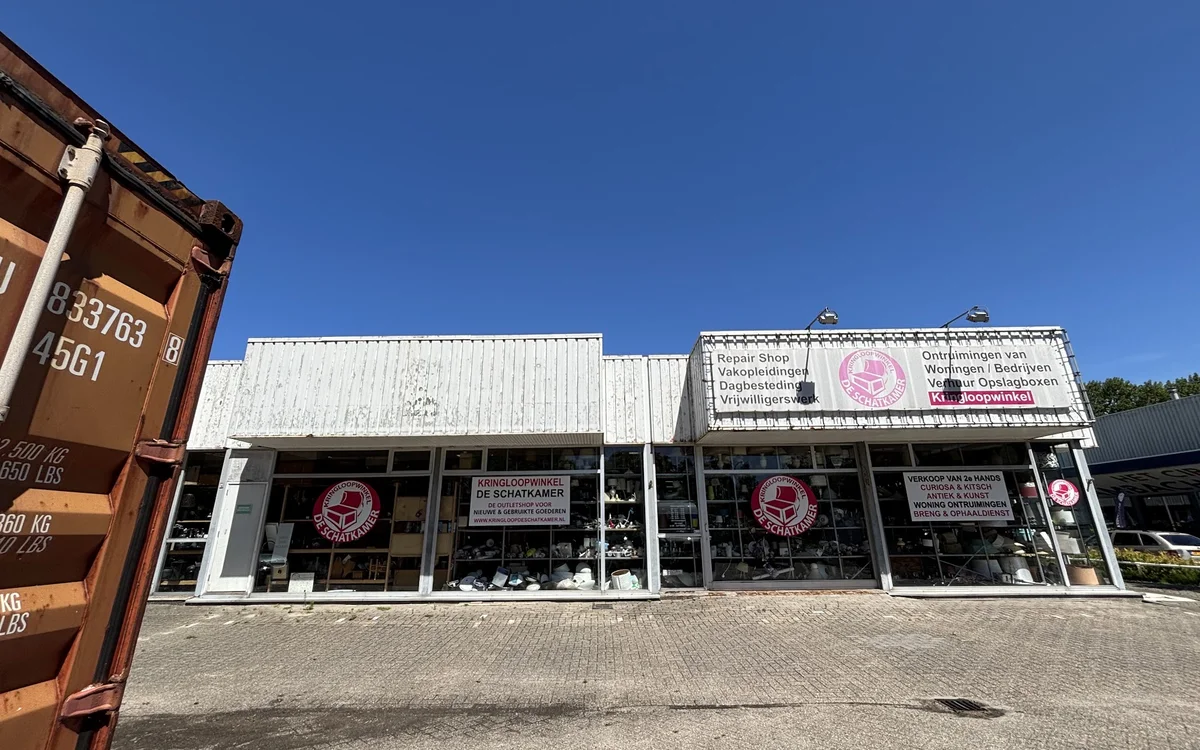 Witte gevel van kringloopwinkel met grote ramen en roze logo's, onder blauwe hemel