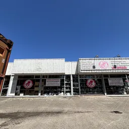 Witte gevel van kringloopwinkel met grote ramen en roze logo's, onder blauwe hemel