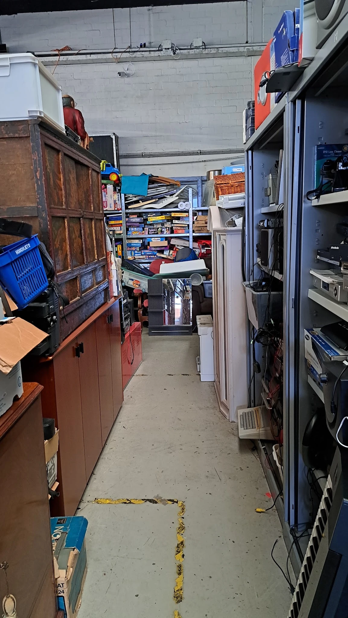Smalle gang in kringloopwinkel met stellingen, dozen en elektronische apparaten aan beide zijden