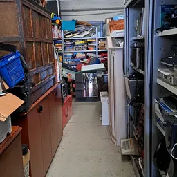 Smalle gang in kringloopwinkel met stellingen, dozen en elektronische apparaten aan beide zijden
