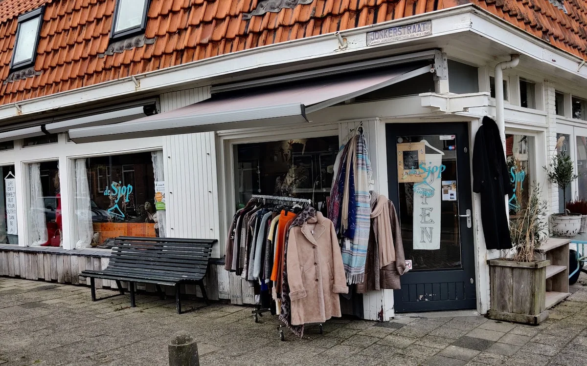 Witte voorgevel van kringloopwinkel met tweedehands kleding uitgestald buiten, rood dak zichtbaar