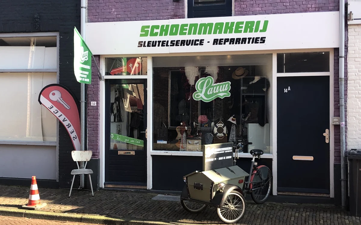 Winkelgevel met groene letters 'SCHOENMAKERIJ' en witte opzetbouw, fietsreparatiewinkel met cargo-fiets op straat