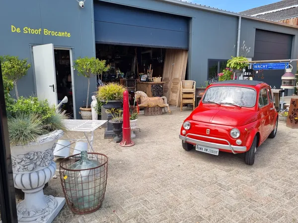 Voorkant van kringloopwinkel met blauwe gevel, grote roldeur, rode vintage auto en plantenbakken