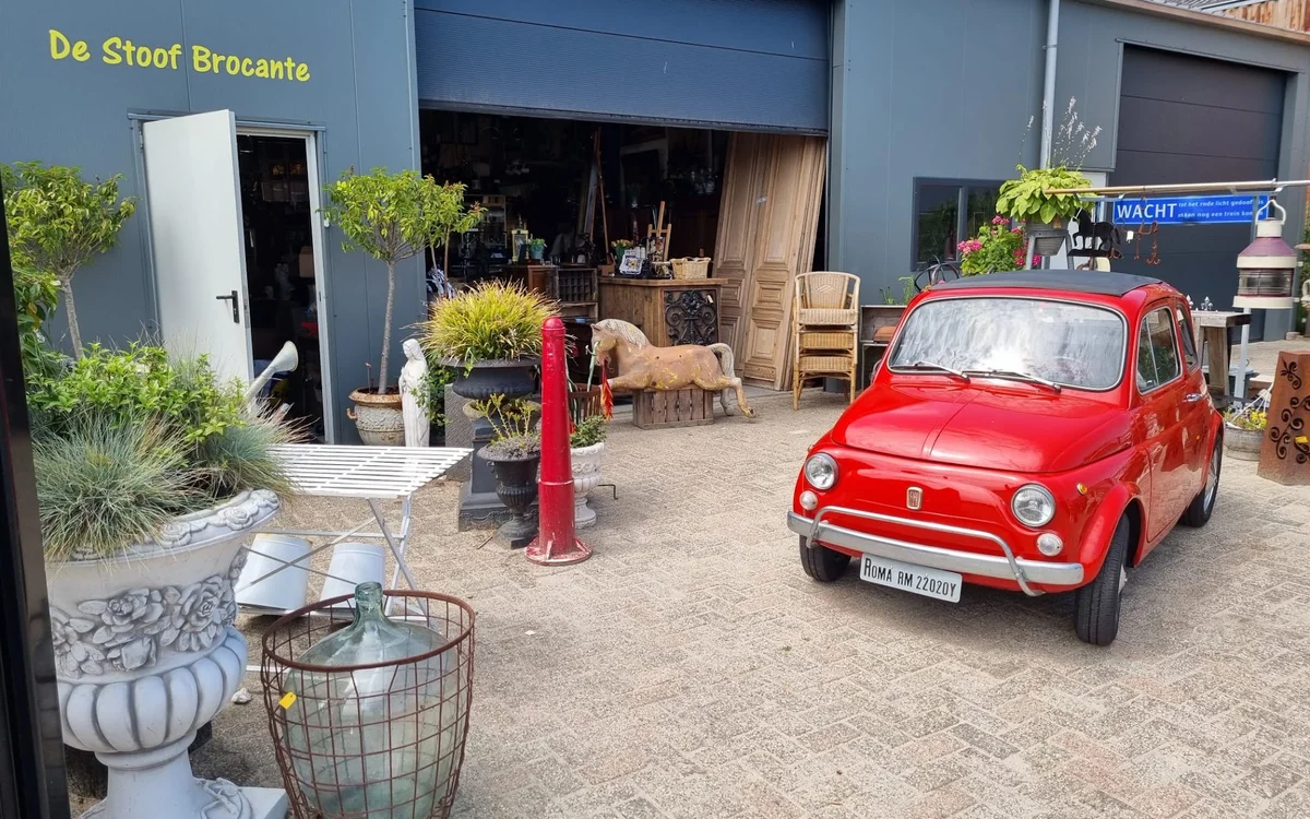 Voorkant van kringloopwinkel De Stoof Brocante met grijze pui, winkelnaam in gele letters, rode vintage auto op het plein, pl
