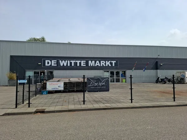 Voorkant van de kringloopwinkel met zwarte gevel en witte letters 'DE WITTE MARKT', omheind parkeerterrein met enkele objecte