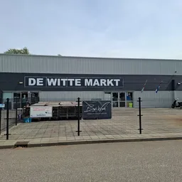 Voorkant van de kringloopwinkel met zwarte gevel en witte letters 'DE WITTE MARKT', omheind parkeerterrein met enkele objecte