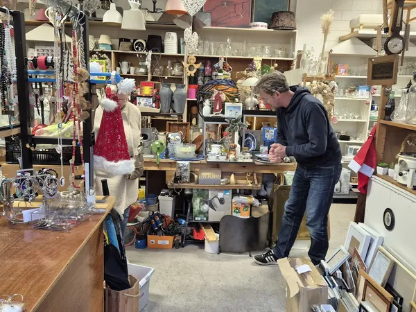 Binnenaanzicht van kringloopwinkel met stellingen vol spullen, etalagepop met rode jurk en man aan het werk