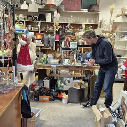 Winkelbinnenruimte met stellingen, diverse spullen, kleding op etalagepop en een medewerker die bezig is in de winkel