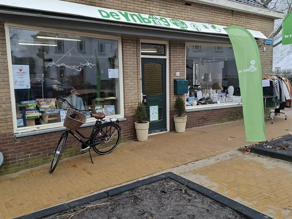 Gevel van kringloopwinkel De Ynbring met groene naam boven, twee etalageruiten met spullen, groene deur en zwarte fiets ervoo