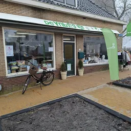 Gevel van kringloopwinkel De Ynbring met groene naam boven, twee etalageruiten met spullen, groene deur en zwarte fiets ervoo