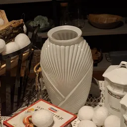 Witte decoratieve vaas met geribfeld oppervlak en touw-detail, omgeven door andere huishoudelijke spullen op een plank.